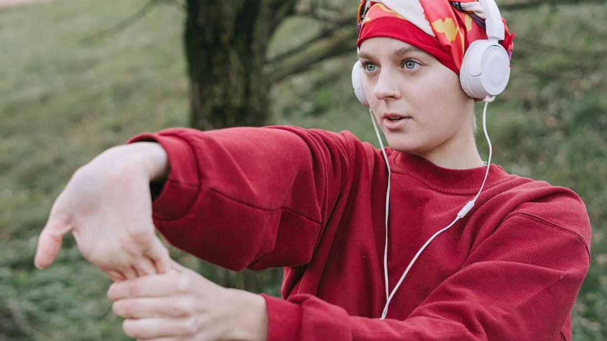 A woman in a red sweater stretches outdoors while listening to music with headphones.