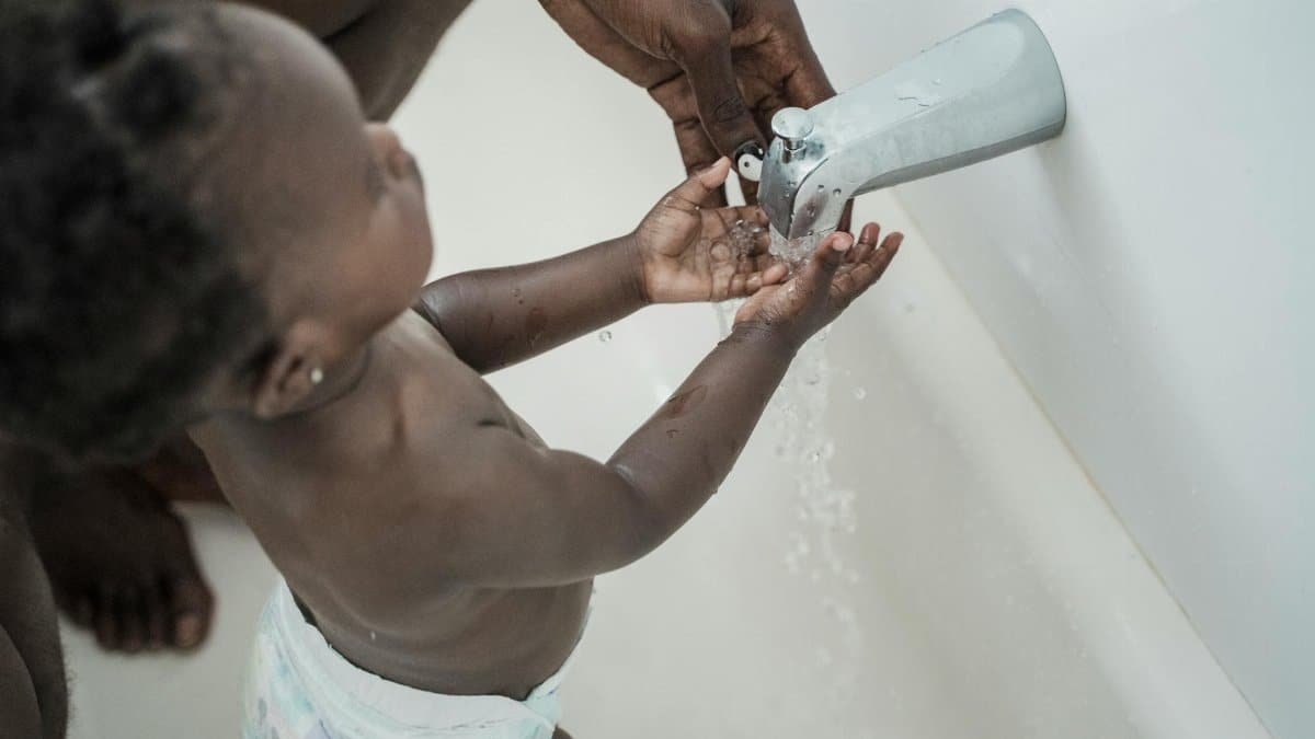 A toddler being guided by an adult to wash hands under a faucet, promoting hygiene.