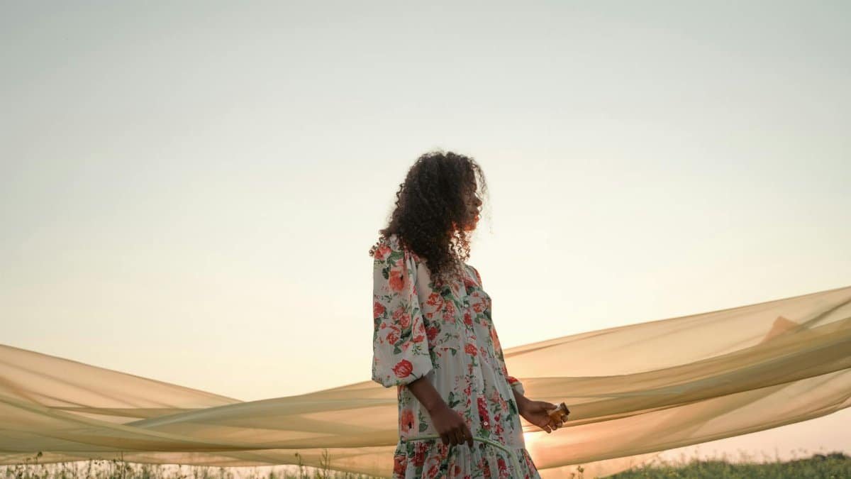 A woman in a floral dress walks gracefully through a sunlit field, with fabric flowing behind her.