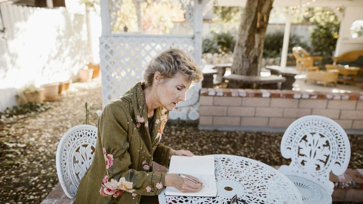 Senior woman writing in a notebook at a garden table, surrounded by nature. Peaceful outdoor scene.