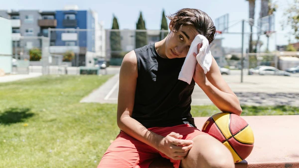 A teenage boy rests on a bench outdoors, wiping sweat after a basketball game.
