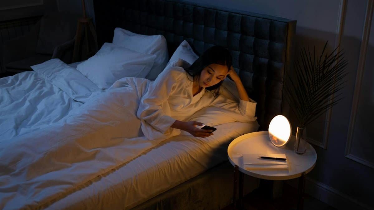 An Asian woman on a bed using her phone in a dim bedroom, reflecting on insomnia or night routines.