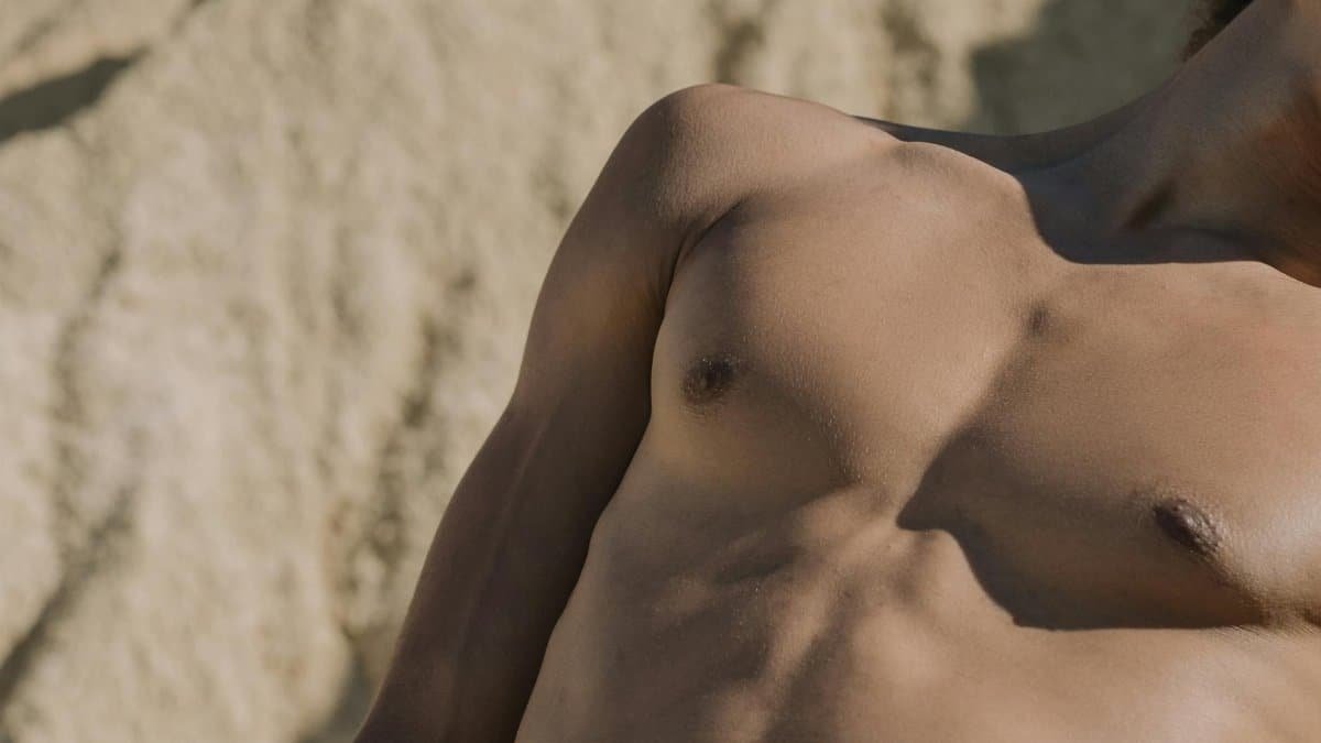 Close-up of a shirtless man's torso with a sandy background.