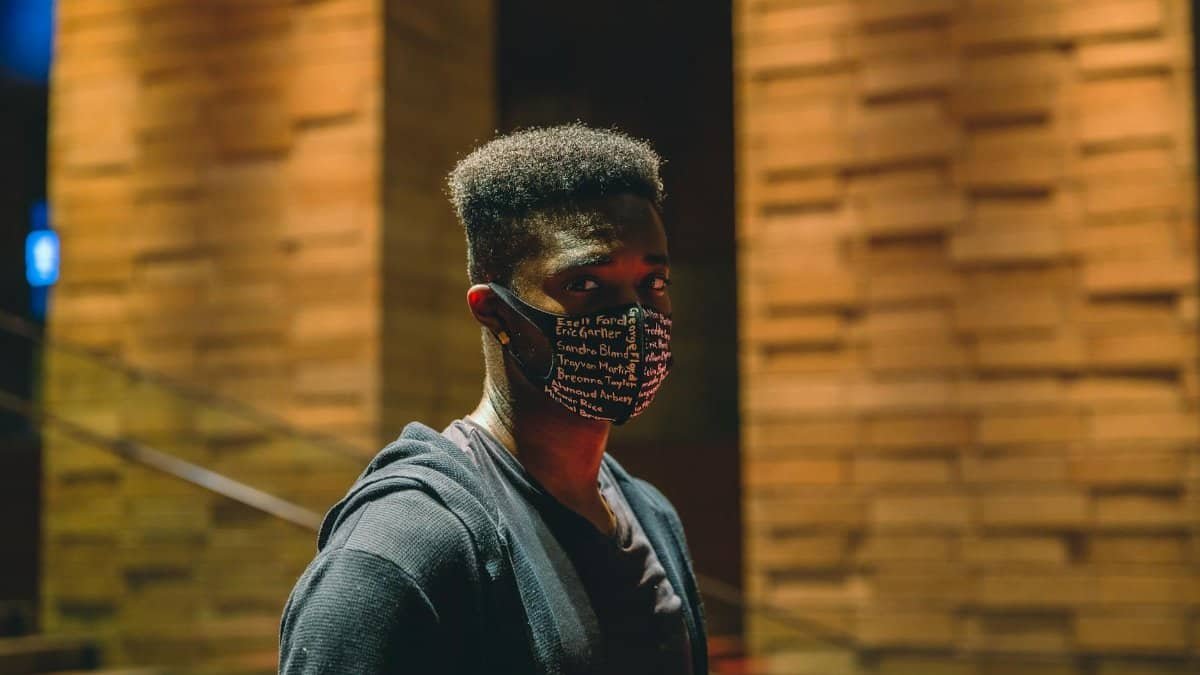 Calm African American male wearing medical mask looking at camera while standing on street near building and staircase in evening time