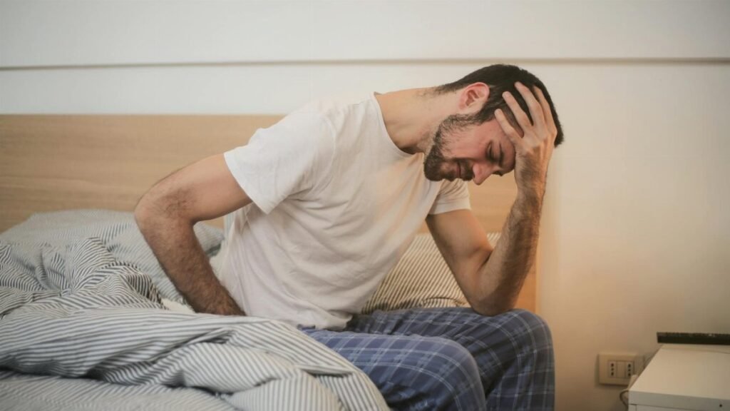A young man in pajamas holding his head, sitting on a bed, appears to be experiencing a headache.