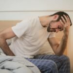 A young man in pajamas holding his head, sitting on a bed, appears to be experiencing a headache.