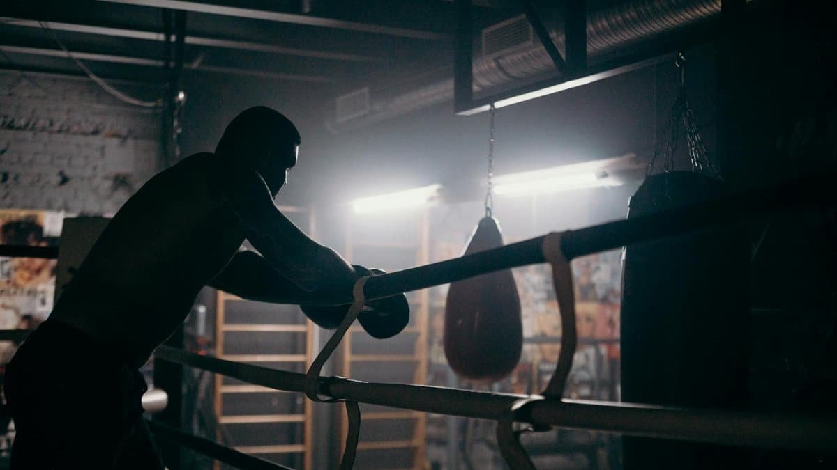 Moody silhouette of a boxer leaning in a dimly lit gym with punching bag and boxing ring.
