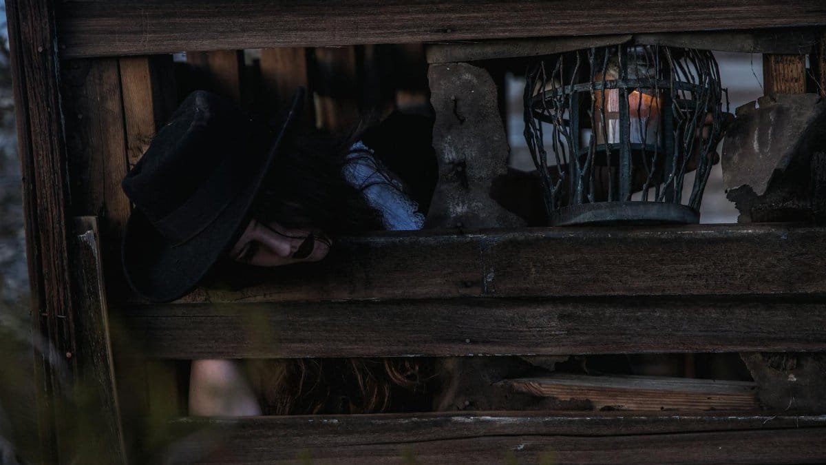 Dark and mysterious scene with a woman in a wooden enclosure, featuring a dimly lit lantern.