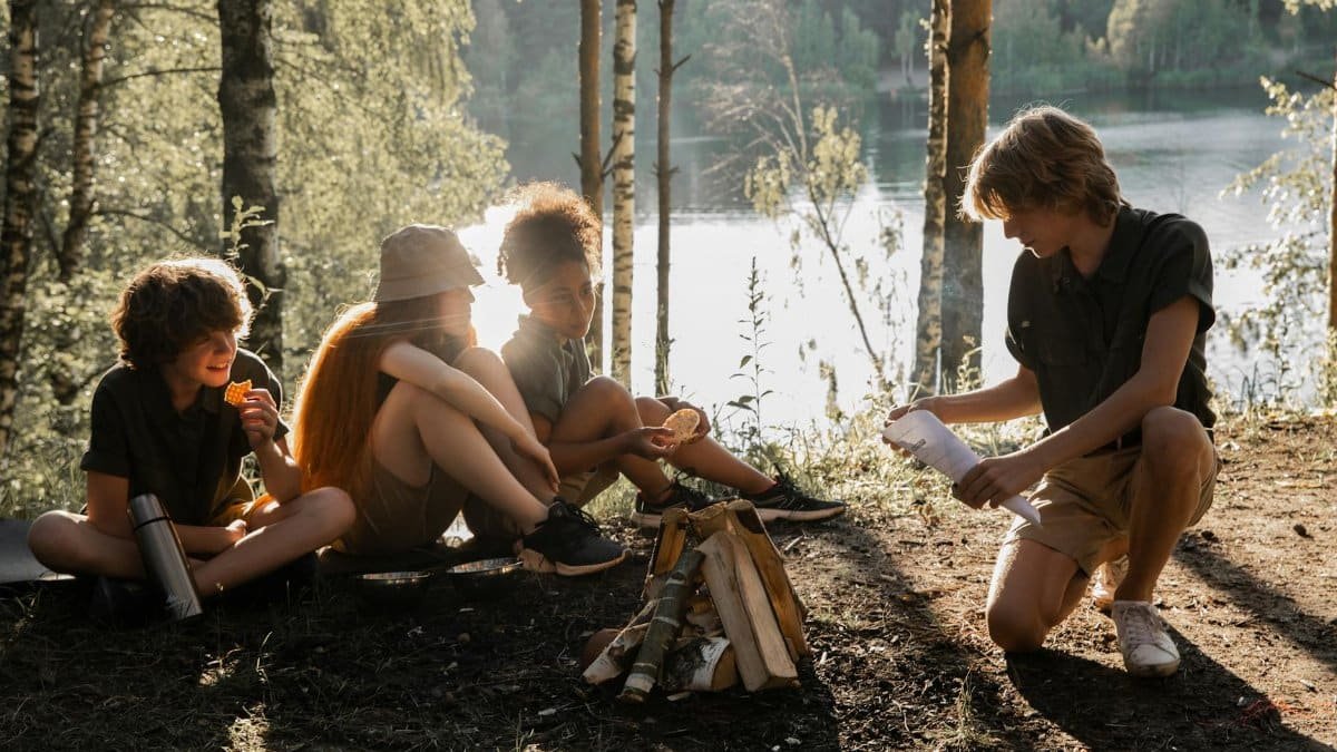 Children enjoying a camping trip by a lake, preparing for a campfire, surrounded by nature at sunset.