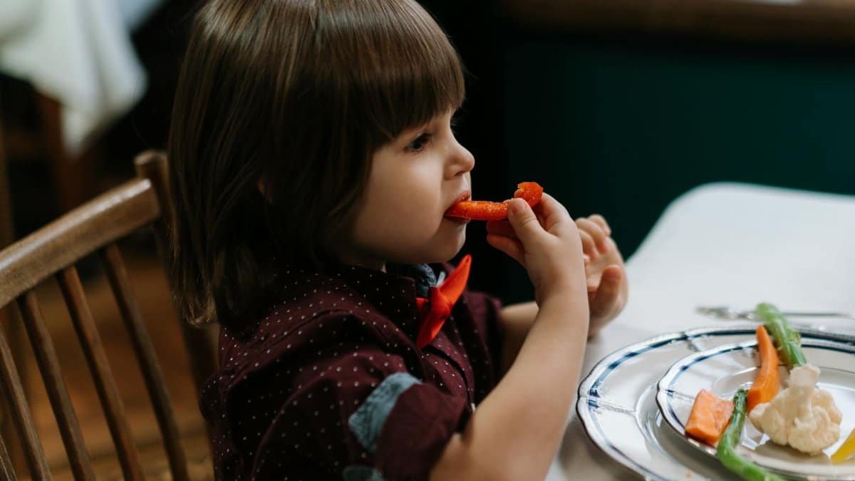 A toddler in a bow tie is eating vegetables such as carrot and cauliflower at a dinner table indoors.