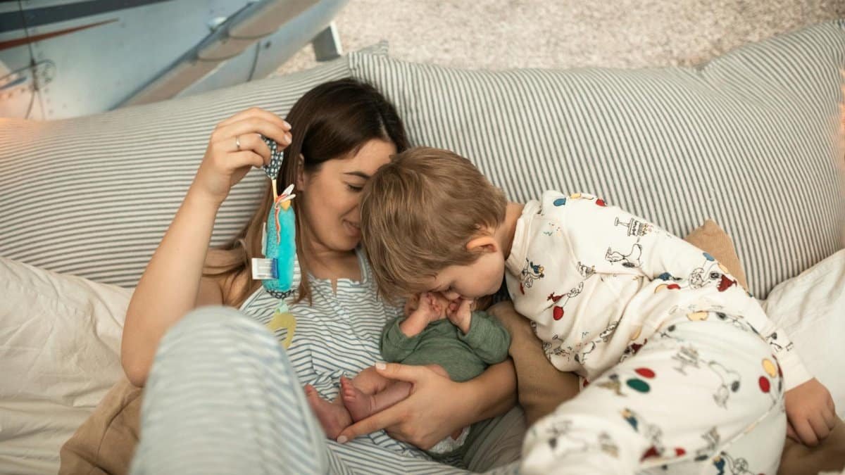 A tender moment captured with a mother, her baby, and older child cuddling in bed.