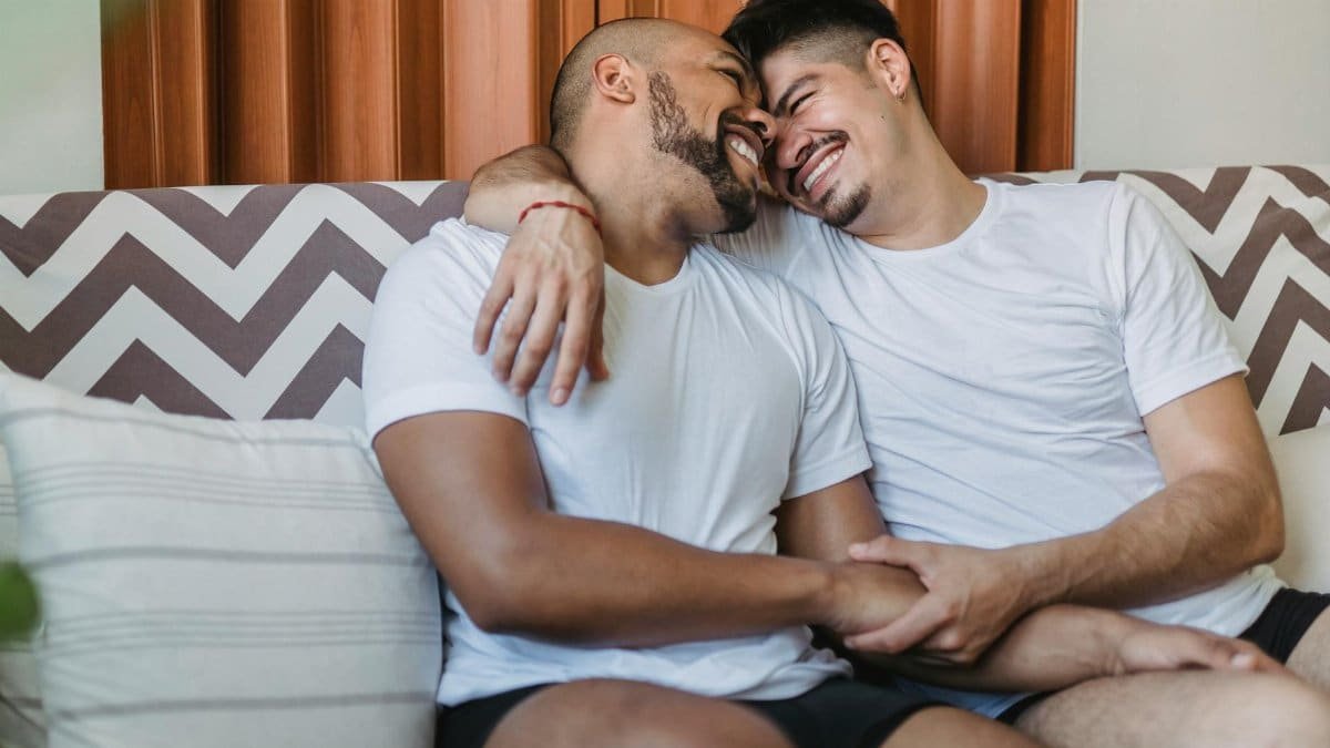 A joyful gay couple cuddling and smiling on a couch, showcasing affection and happiness.