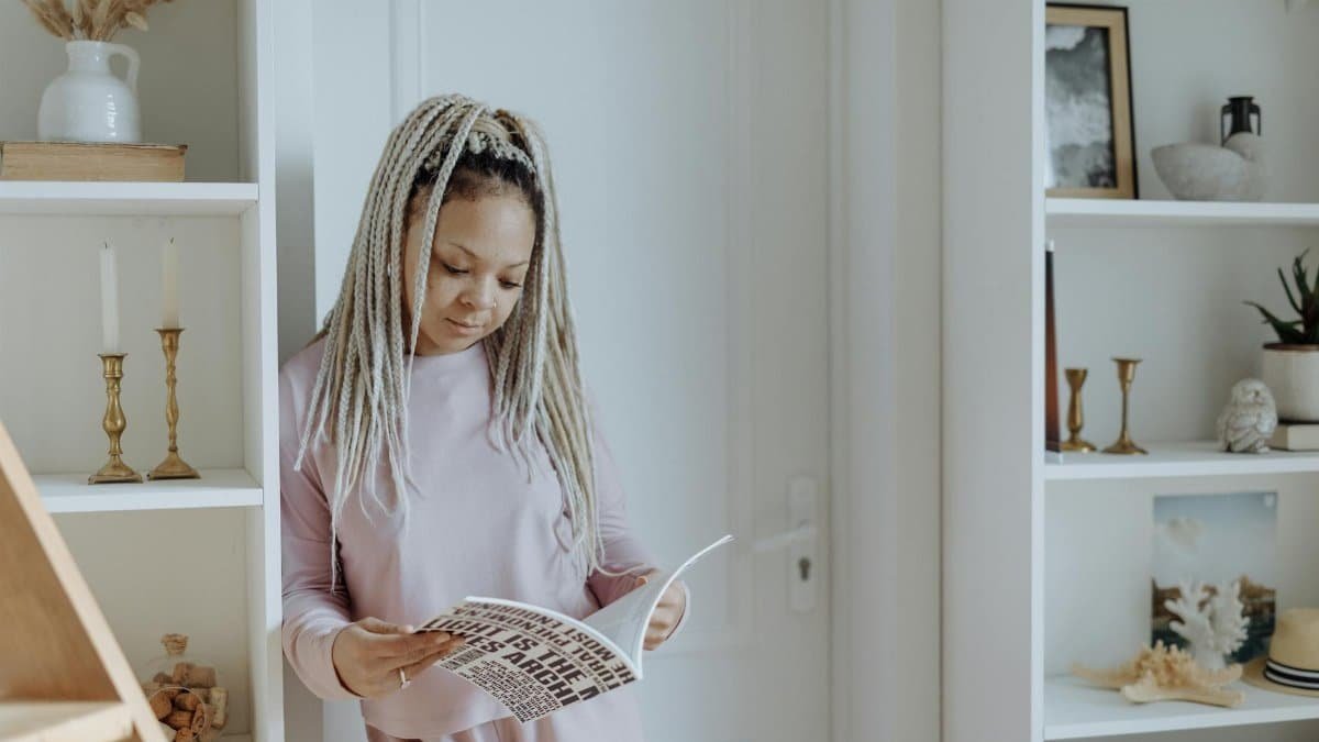 Black woman with long braids reading in stylish home interior, against white background.