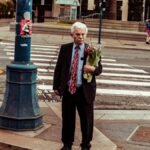 Elderly man holding flowers at a San Francisco crosswalk, vibrant urban scene.