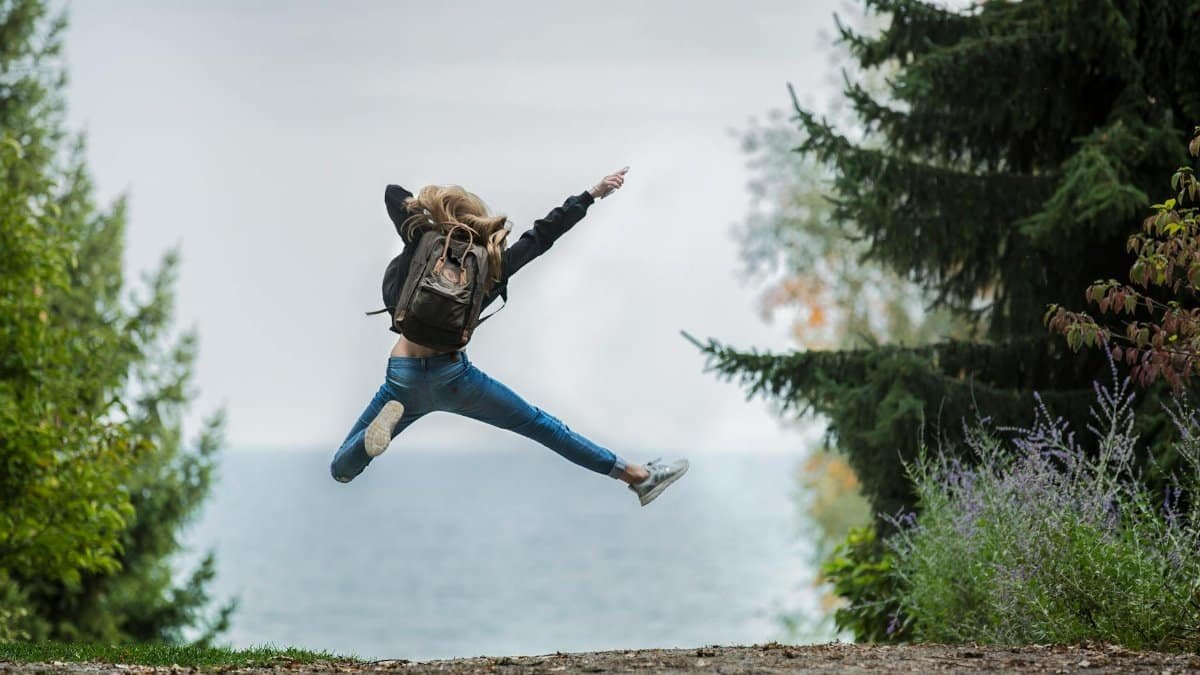 Energetic woman leaping in outdoor forest setting with lake view, showcasing freedom and joy.