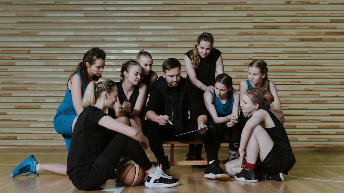 A coach strategizes with a women's basketball team in a gymnasium.