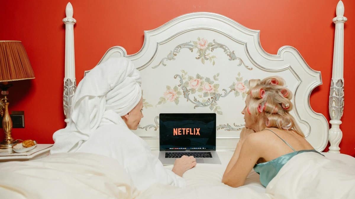 Two women lying in bed watching Netflix on a laptop, creating a cozy and bonding atmosphere.