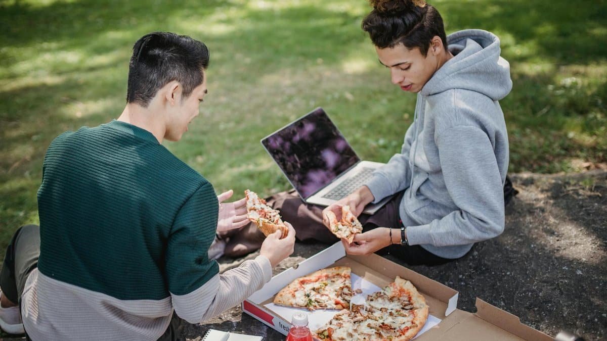 Two friends relaxing outdoors with pizza and a laptop on a sunny day.