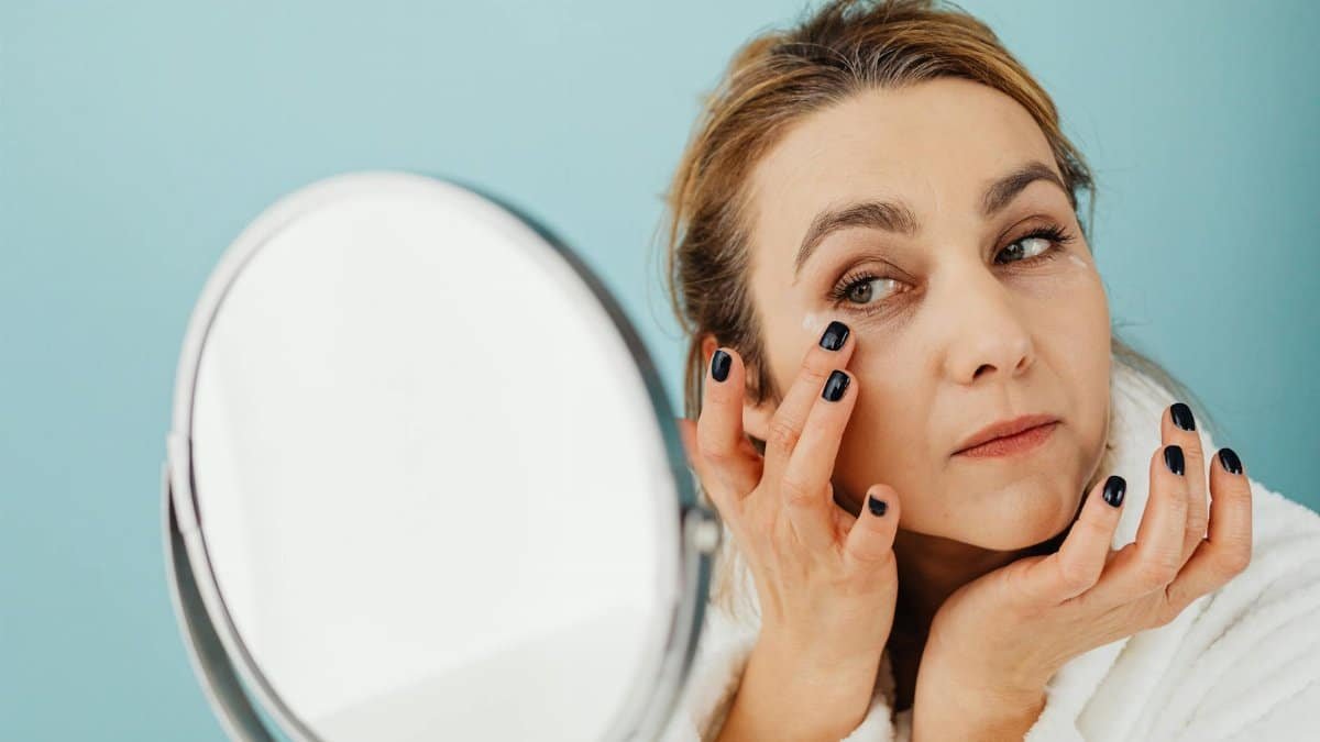 A woman in a bathrobe applies skincare cream while looking in the mirror. Ideal for beauty and self-care concepts.