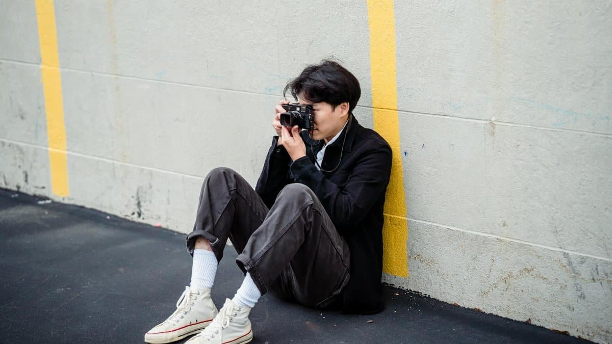 A candid shot of a man sitting against a wall photographing outdoors.