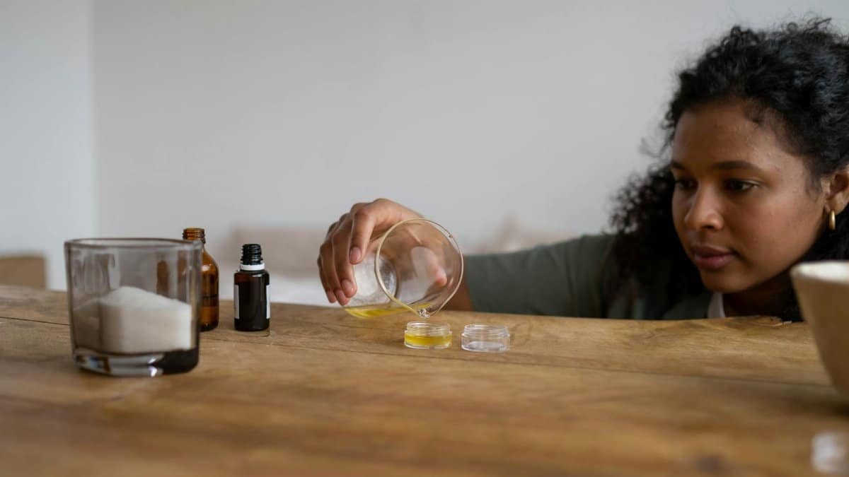 A woman mixes ingredients to create natural lip balm indoors, using a wooden table as a workspace.