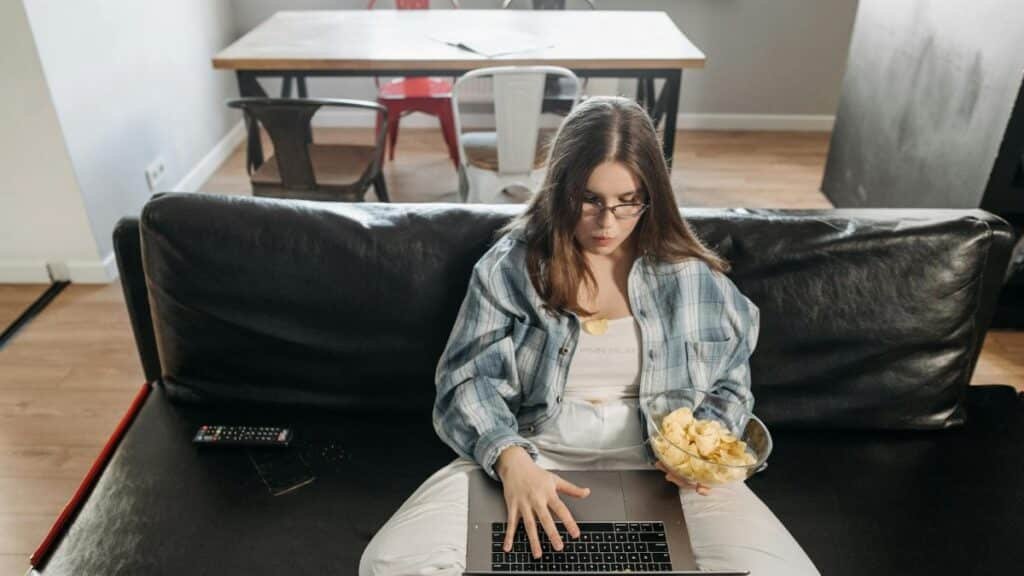 Young woman sitting on couch with laptop and snacks, working from home in a modern living room.