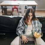 Young woman sitting on couch with laptop and snacks, working from home in a modern living room.