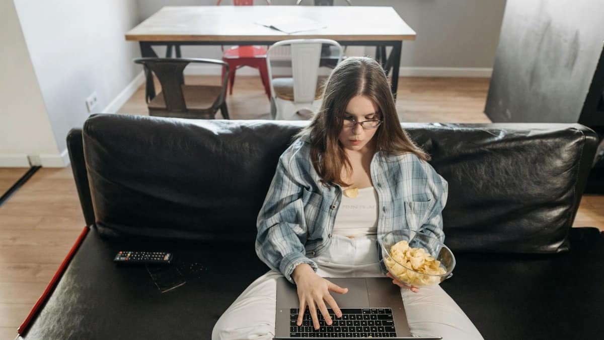 Young woman sitting on couch with laptop and snacks, working from home in a modern living room.