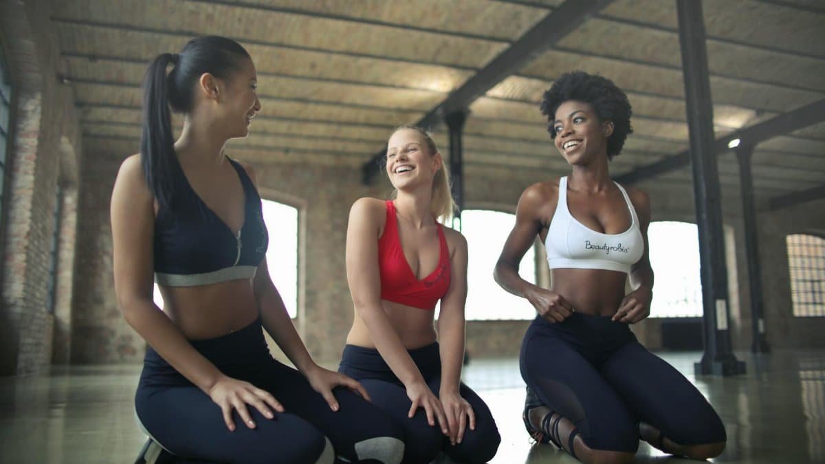 Three women in activewear laughing and exercising together indoors in a gym setting.