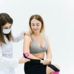 A therapist applies kinesio tape on a woman's knee during a physical therapy session on a white background.