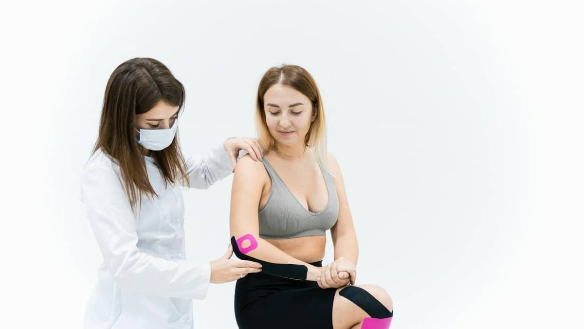 A therapist applies kinesio tape on a woman's knee during a physical therapy session on a white background.