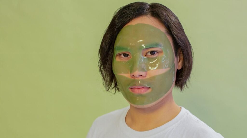 Adult wearing a green facial mask on a light green background, promoting skincare and wellness.