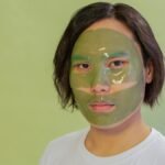 Adult wearing a green facial mask on a light green background, promoting skincare and wellness.