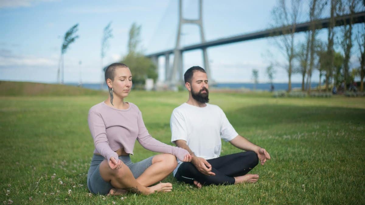 A couple practices meditation outdoors, embodying relaxation and mindfulness beneath the Vasco da Gama Bridge, Lisbon.