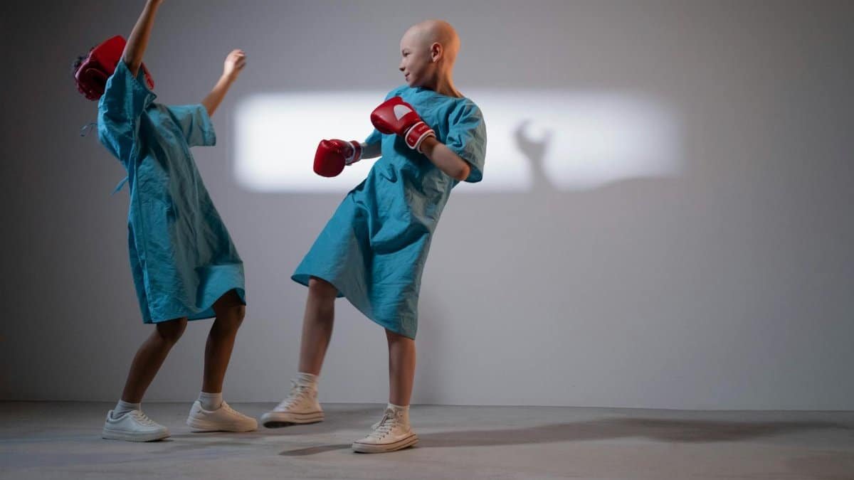 Two children in hospital gowns engage in playful boxing in a well-lit room.