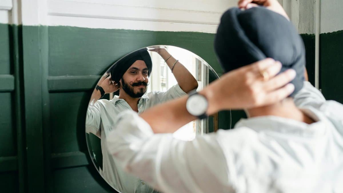 Back view of satisfied young Indian male hipster with twisted mustache wearing denim shirt and turban standing against mirror on wall while taking care about appearance