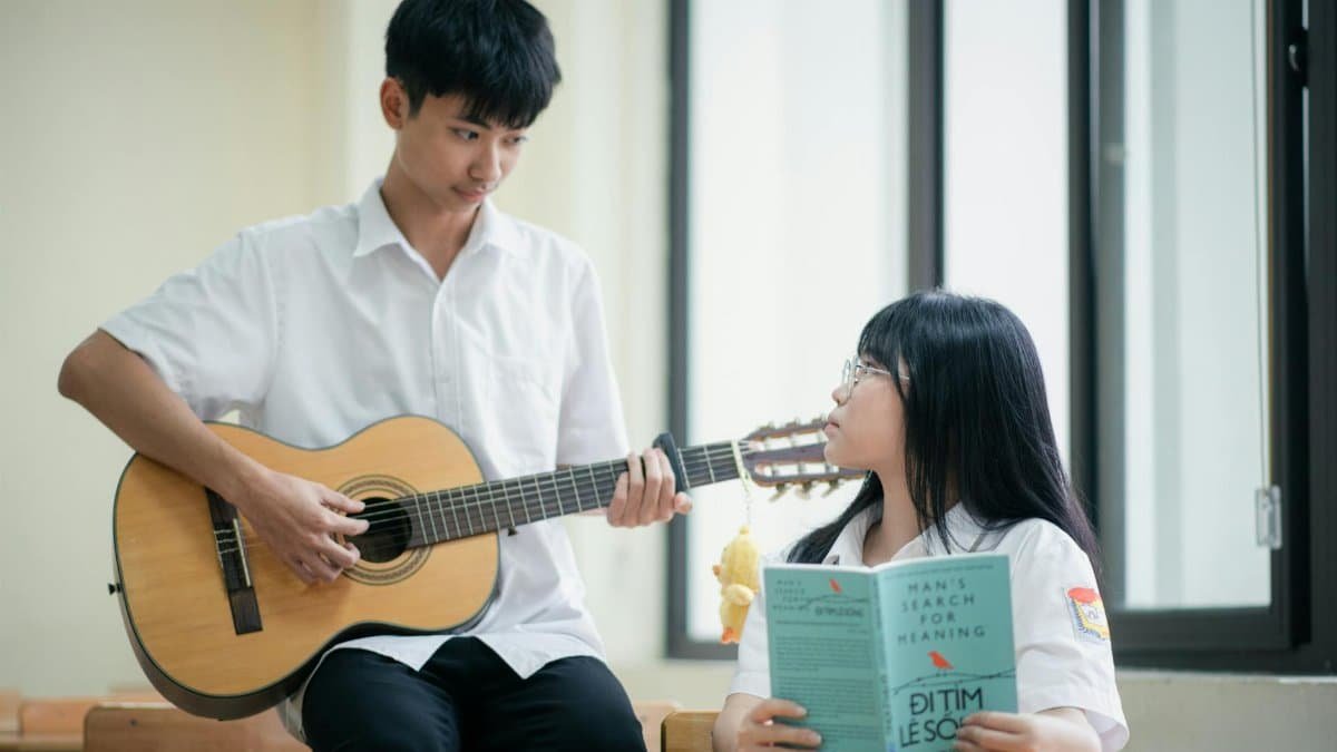 Two teenagers in a classroom: one playing guitar, another reading, engaging in a learning atmosphere.