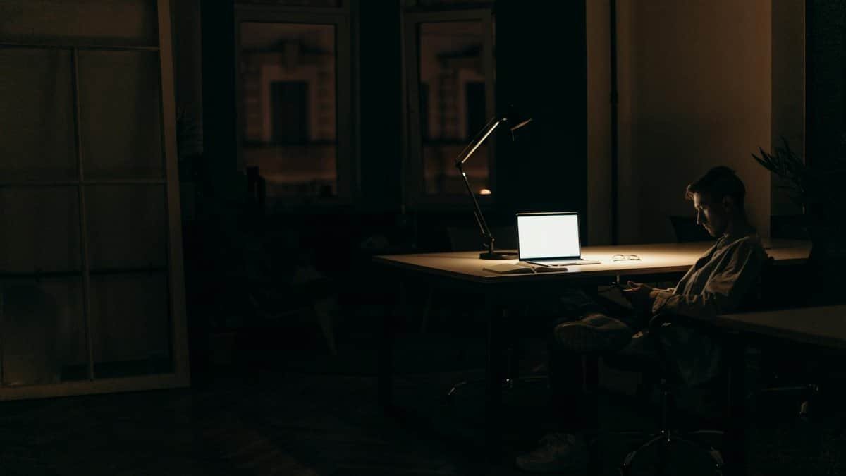 A man working late at night in a dimly lit office, with a bright laptop screen.