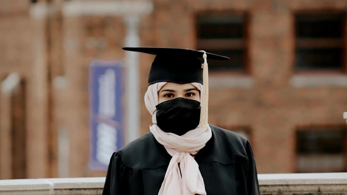 Female graduate with hijab and face mask standing outdoors, emphasizing education and resilience.