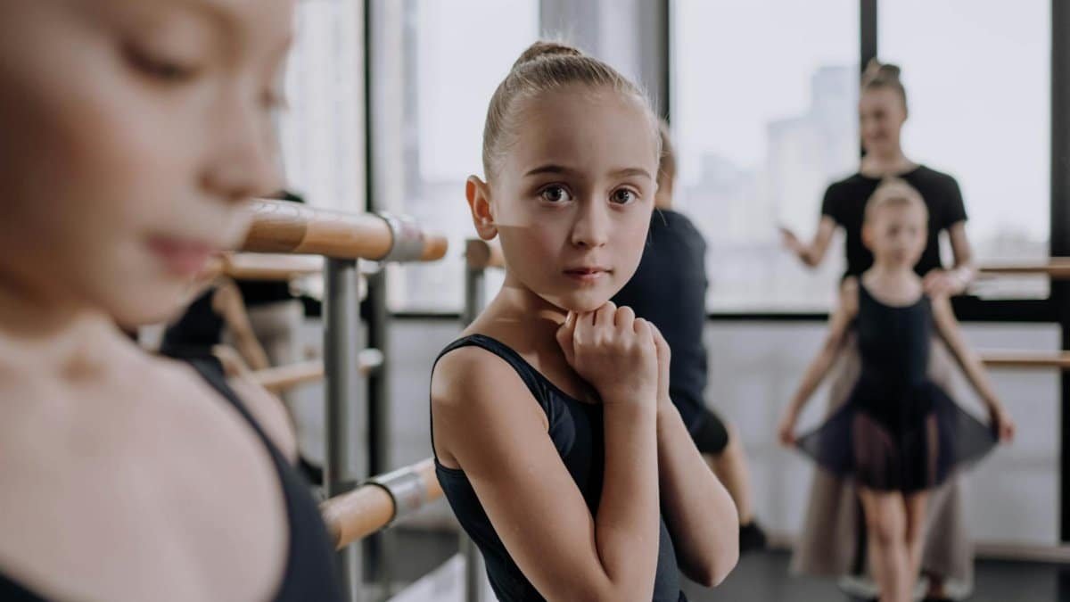 A group of children learning ballet in a studio with natural light, practicing at the barre.