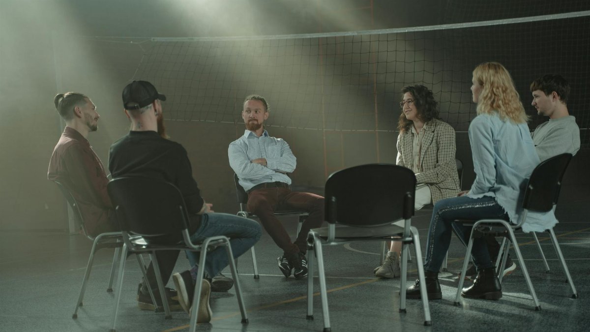 A group therapy session with six adults seated in a circle, discussing support and mental health.