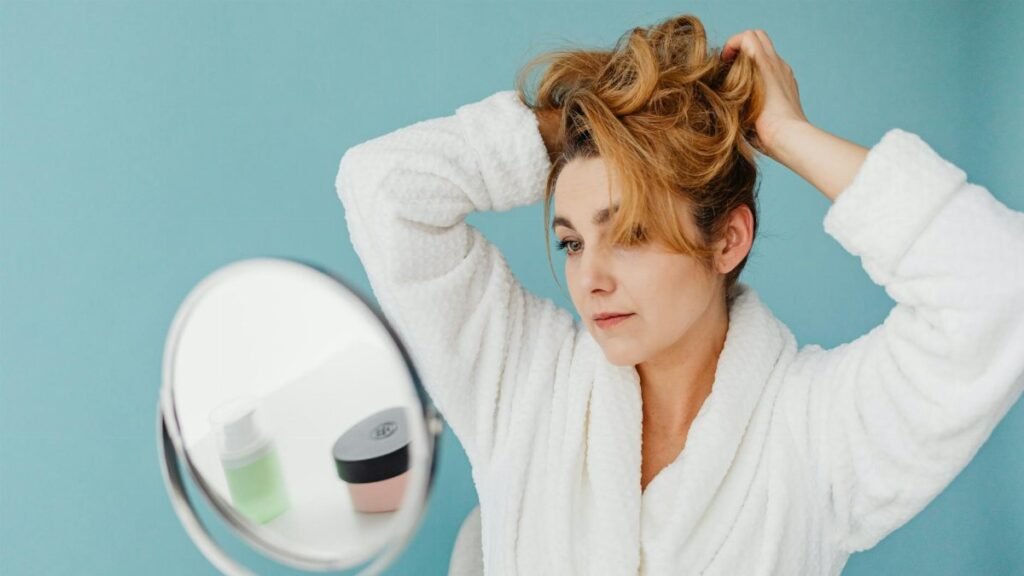 Woman in bathrobe looks in mirror while styling hair, in a modern indoor setting.