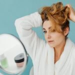 Woman in bathrobe looks in mirror while styling hair, in a modern indoor setting.
