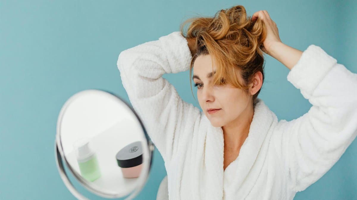 Woman in bathrobe looks in mirror while styling hair, in a modern indoor setting.