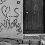 A cat sits on steps beside a graffiti-covered wall with a wooden door, captured in black and white.