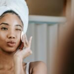 Charming ethnic female with folded lips winking while cleaning face with cotton pad during daily beauty routine