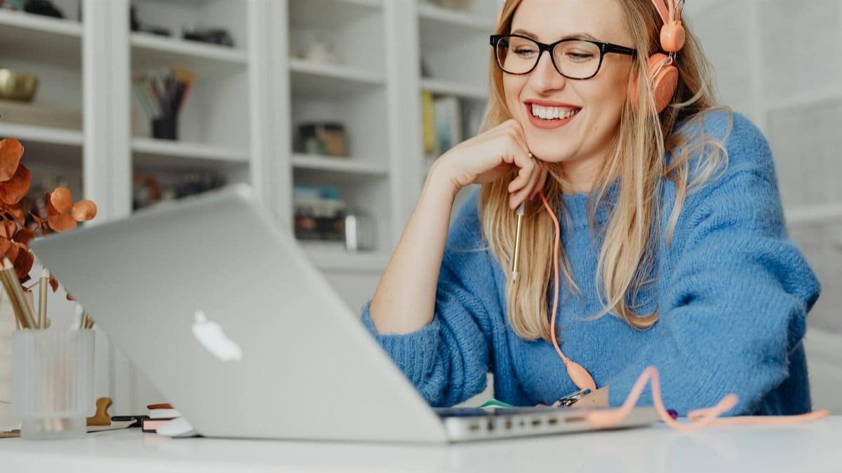 Smiling woman wearing headphones and glasses, working on a laptop in a bright indoor setting.