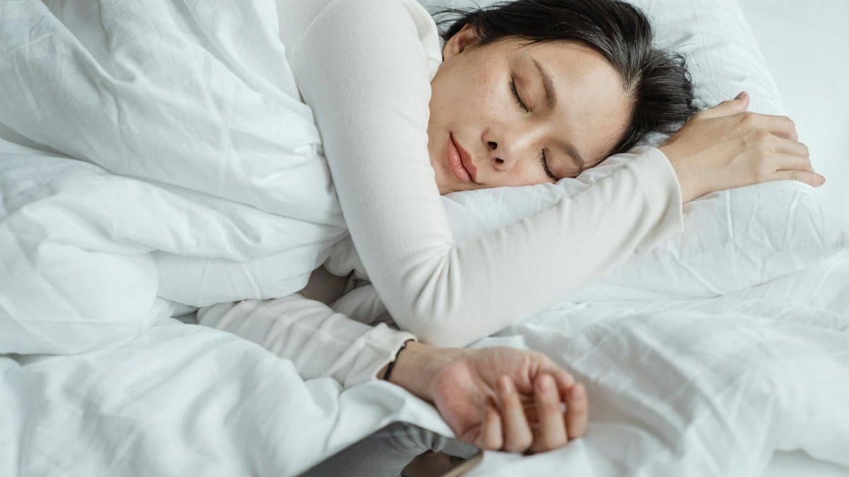 Calm Asian female wearing white pajama sleeping in comfortable bed with white sheets near modern mobile phone in morning