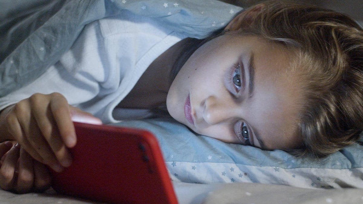 Child lying in bed under a blanket, focused on a smartphone screen, displaying a bedtime routine.