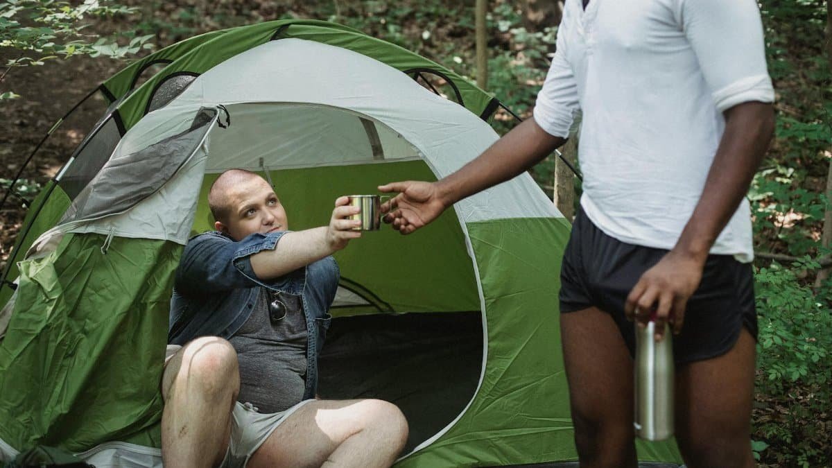 Two young men camping, sharing a warm drink from a thermos in a forest setting.
